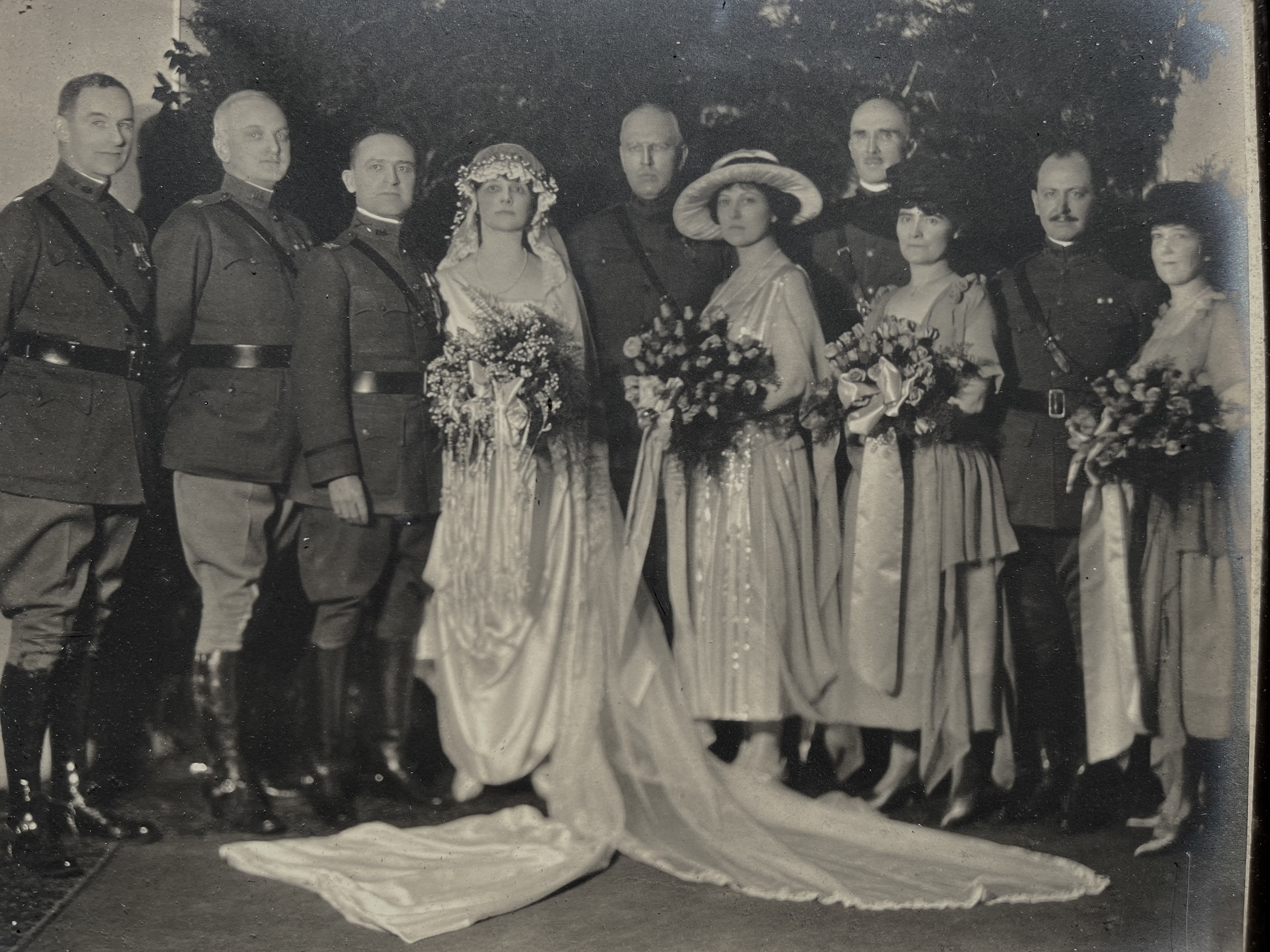 1921 photo of Perry wedding party, with male members in uniforms and females holding flowers against floral backdrop.